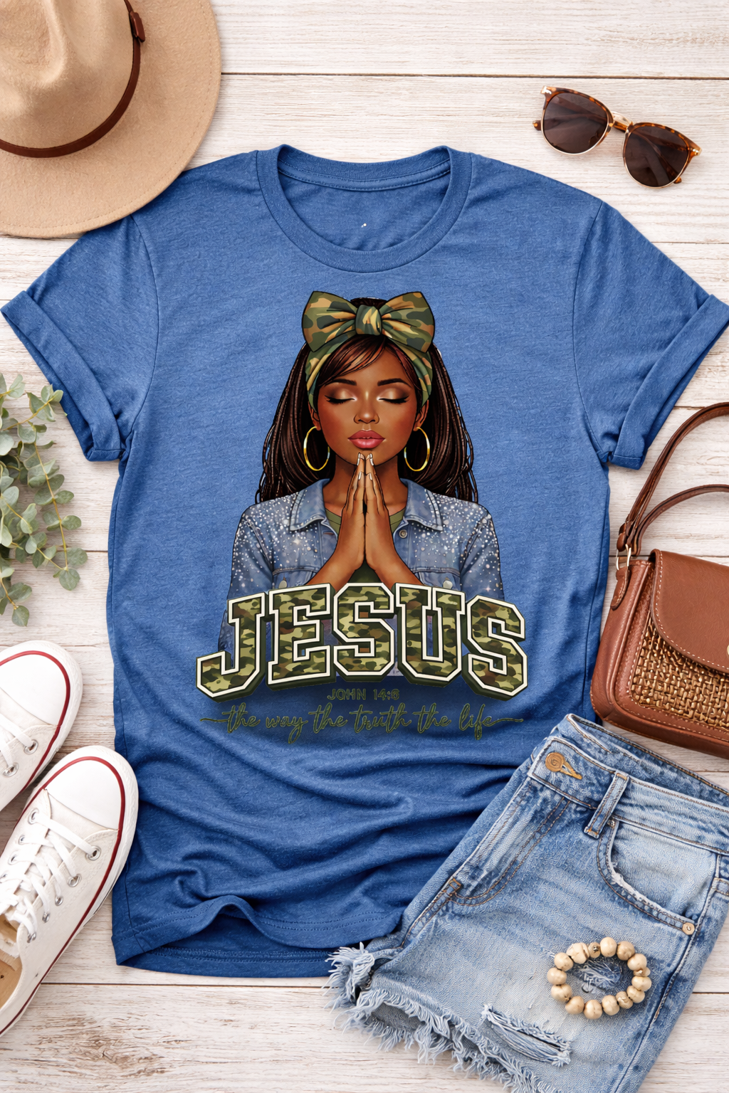 Blue t-shirt with a graphic of a woman with a bow and 'Jesus' text, surrounded by casual clothing items on a wooden surface.
