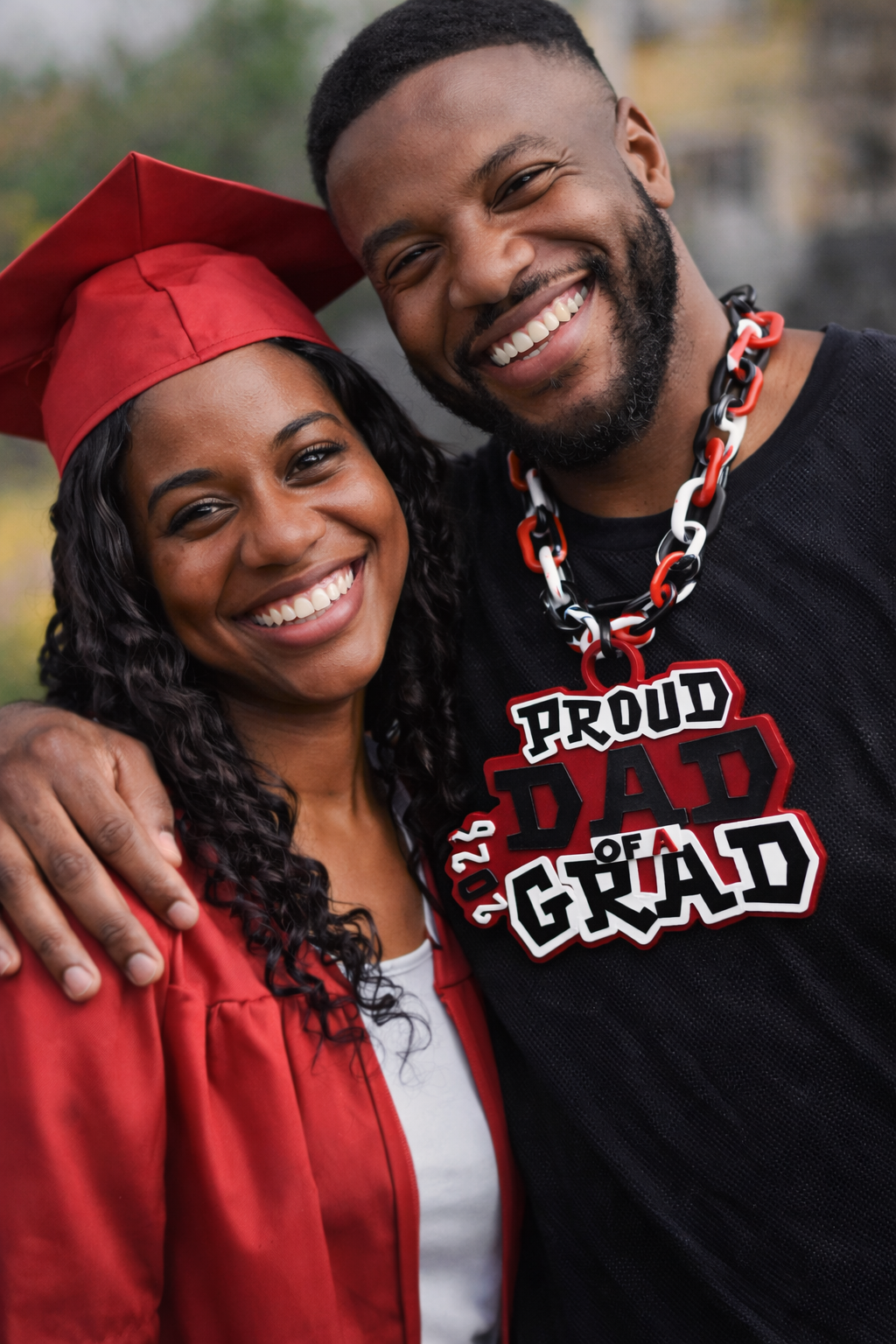 Graduating student in red cap and gown with proud father wearing 'Proud Dad of a Grad' shirt.